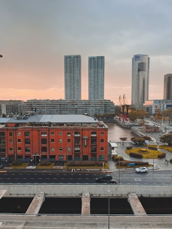 A cityscape featuring a tall red-brick building in the foreground with large windows and a modern roof. Behind it, there are two sleek, high-rise apartment buildings and another tower with the letters YPF visible at the top. The scene includes a river or canal with a dock and cranes. The sky is a mix of grey and soft orange tones, suggesting either dawn or dusk. Vehicles are visible on the road in front of the buildings, and there are carefully maintained lawns and pathways alongside the river.