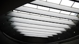 A modern architectural ceiling with large, rectangular skylights arranged in parallel lines, providing natural light to the interior space. The ceiling is bordered by dark walls, and multiple small lights are suspended in between the skylights.