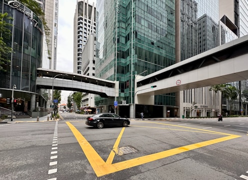 A modern cityscape features tall glass buildings and a pedestrian bridge connecting structures above the street. The intersection has yellow grid lines and a few vehicles, including a black car and a motorcycle. Sidewalks are bordered by greenery, and the area appears to be an urban business district.