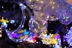 A cluster of glowing spherical light bulbs with delicate wire-like structures inside. A crescent moon-shaped decoration is wrapped in string lights, adding a dreamy ambiance. The background is filled with a vibrant array of colored lights, creating a festive atmosphere.