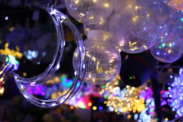 A cluster of glowing spherical light bulbs with delicate wire-like structures inside. A crescent moon-shaped decoration is wrapped in string lights, adding a dreamy ambiance. The background is filled with a vibrant array of colored lights, creating a festive atmosphere.