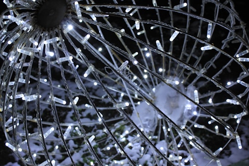 A close-up view of a spherical wire structure adorned with numerous small, white LED lights. The sphere appears to be part of a decorative outdoor installation, glowing brightly against a dark background. Snow is visible on some of the structure, enhancing the wintry atmosphere.