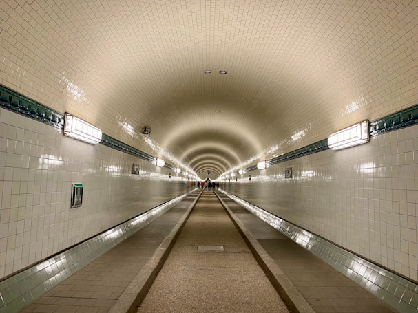A long, tiled underground tunnel with an arched ceiling that features evenly spaced light fixtures. The tunnel is lined with a narrow walkway and wider path in the middle. The lighting creates a warm atmosphere and a vanishing point at the tunnel's end where a few people are walking.