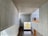 A modern architectural interior featuring tall concrete walls with a large, rectangular window letting in natural light. The space has minimalist design elements with clean lines and large, open areas. A person stands at the far end near the window, providing a sense of scale.