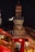 A domed clock tower is illuminated at night with a crescent moon visible in the sky. Below the tower, the inscription reads 'Centro Canalejas' and features golden decorative elements with sculptures. In the foreground, a red vehicle with reflections of festive lights is visible, and a lit Christmas tree can be seen in the background.