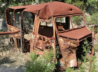 A heavily rusted and dilapidated vehicle sits in an outdoor setting surrounded by greenery. The vehicle appears abandoned and is missing significant parts, with its metal frame showing extensive corrosion.