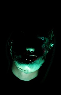 A dark and eerie image featuring a partially illuminated face with a hood. The face has an unnatural, shadowy appearance with green and subtle red highlights, giving it a mysterious and unsettling vibe.