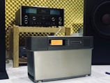 A vintage audio setup featuring an amplifier on a wooden shelf and a speaker system positioned in front. The background consists of acoustic foam panels, and a partial view of a large speaker is visible to the right.