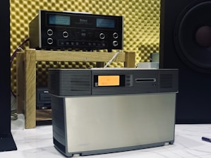 A vintage audio setup featuring an amplifier on a wooden shelf and a speaker system positioned in front. The background consists of acoustic foam panels, and a partial view of a large speaker is visible to the right.