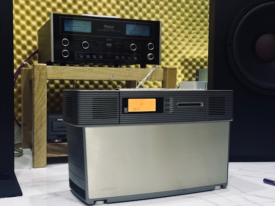 A vintage audio setup featuring an amplifier on a wooden shelf and a speaker system positioned in front. The background consists of acoustic foam panels, and a partial view of a large speaker is visible to the right.