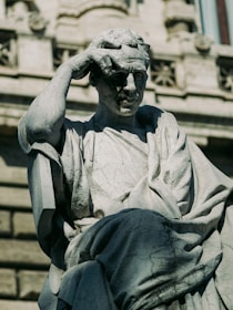 A stone statue depicting a contemplative figure with one hand on its head, possibly in thought. The figure wears draped clothing and is set against a classical architectural background, with intricate detailing and a window visible in the upper portion.
