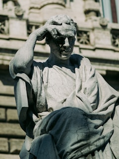 A stone statue depicting a contemplative figure with one hand on its head, possibly in thought. The figure wears draped clothing and is set against a classical architectural background, with intricate detailing and a window visible in the upper portion.