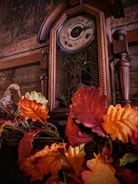 An antique wooden clock with intricate designs stands adorned by vibrant autumn leaves. The clock face shows Roman numerals, and the background features a vintage mural with faded text and images.