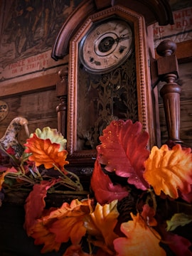 An antique wooden clock with intricate designs stands adorned by vibrant autumn leaves. The clock face shows Roman numerals, and the background features a vintage mural with faded text and images.