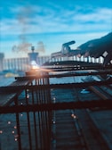 A person is welding metal bars with bright sparks flying around. The setting appears industrial, with a hazy background and a blue sky. The focus is on the welding process and the tools in hand.