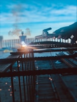 A person is welding metal bars with bright sparks flying around. The setting appears industrial, with a hazy background and a blue sky. The focus is on the welding process and the tools in hand.