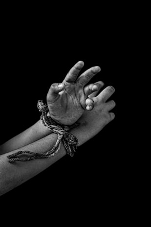 A pair of hands bound together with a rope, positioned against a dark background. The hands appear to be dirty or smudged, emphasizing a sense of struggle or distress.