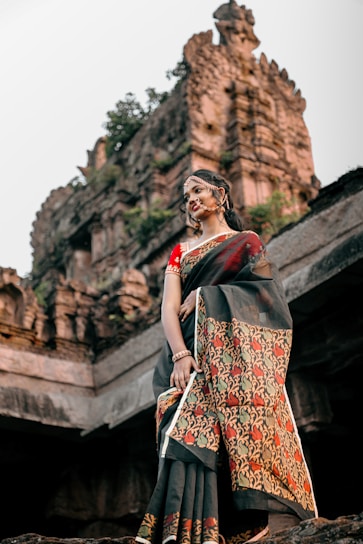 A graceful woman draped in a richly embroidered traditional saree, standing against a backdrop of Indian architecture.