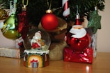 A variety of Christmas ornaments are displayed, including a snow globe with Santa Claus inside, a red bauble with a Santa face, and several glittery green, red, and gold ornaments. These decorations hang from a Christmas tree that is partly visible, alongside gift boxes wrapped in shiny paper.