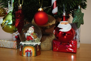 A variety of Christmas ornaments are displayed, including a snow globe with Santa Claus inside, a red bauble with a Santa face, and several glittery green, red, and gold ornaments. These decorations hang from a Christmas tree that is partly visible, alongside gift boxes wrapped in shiny paper.
