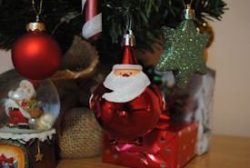 A Christmas-themed scene features various decorations including a red spherical ornament with a Santa face, a glittery green star ornament, a candy cane with red and white stripes, and a Santa figurine inside a snow globe. Burlap and wrapped gifts add to the festive atmosphere.