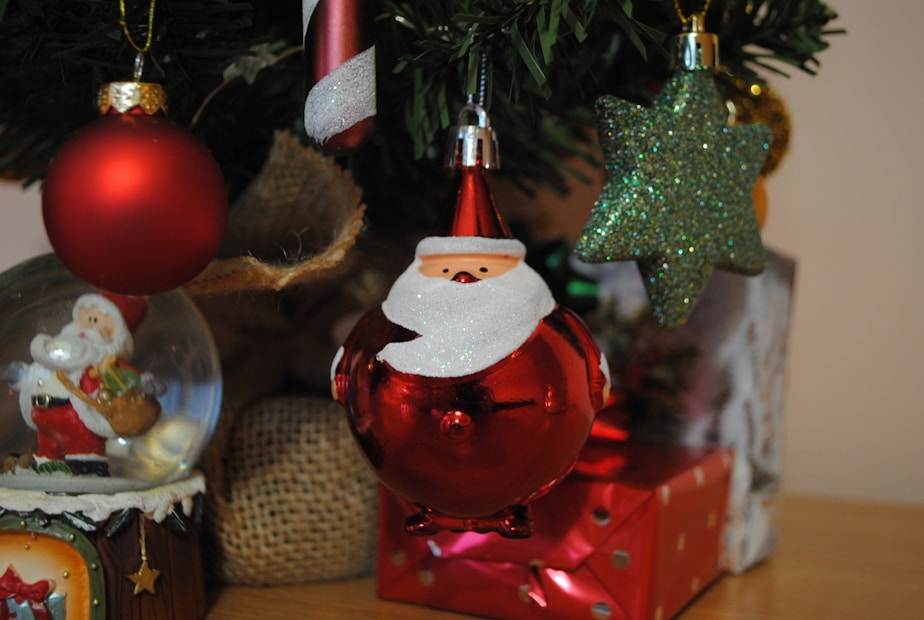 A Christmas-themed scene features various decorations including a red spherical ornament with a Santa face, a glittery green star ornament, a candy cane with red and white stripes, and a Santa figurine inside a snow globe. Burlap and wrapped gifts add to the festive atmosphere.