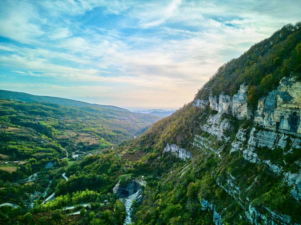 The image presents a breathtaking view of a valley nestled between two towering cliffs, with a river meandering through it. The valley is lush and green, dotted with trees that add to its natural beauty. The sky above is a clear blue, providing a striking contrast to the verdant landscape below. The perspective of the photo is from an elevated vantage point, offering a panoramic view of this picturesque scene.

The image does not contain any discernible text or countable objects, and there are no people present in the frame.