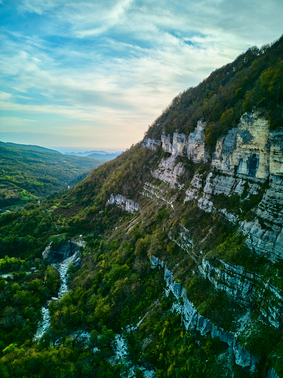 The image presents an aerial view of a mountainous landscape, with a river meandering through it. The river is surrounded by lush green vegetation and rocky cliffs on both sides. The sky above is a clear blue, dotted with fluffy white clouds. The perspective from the top of the mountain provides a comprehensive view of the valley below, showcasing the natural beauty of the region.