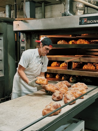 A baker wearing a white uniform and a red cap is carefully removing freshly baked bread from an industrial oven. The bread is displayed on a conveyor and appears golden brown with a dusting of flour on top. The setting is a commercial bakery with stainless steel equipment and machinery visible in the background.