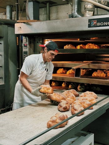 A baker wearing a white uniform and a red cap is carefully removing freshly baked bread from an industrial oven. The bread is displayed on a conveyor and appears golden brown with a dusting of flour on top. The setting is a commercial bakery with stainless steel equipment and machinery visible in the background.
