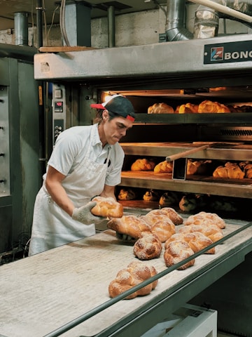 A baker wearing a white uniform and a red cap is carefully removing freshly baked bread from an industrial oven. The bread is displayed on a conveyor and appears golden brown with a dusting of flour on top. The setting is a commercial bakery with stainless steel equipment and machinery visible in the background.