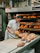 A baker wearing a white uniform and a red cap is carefully removing freshly baked bread from an industrial oven. The bread is displayed on a conveyor and appears golden brown with a dusting of flour on top. The setting is a commercial bakery with stainless steel equipment and machinery visible in the background.
