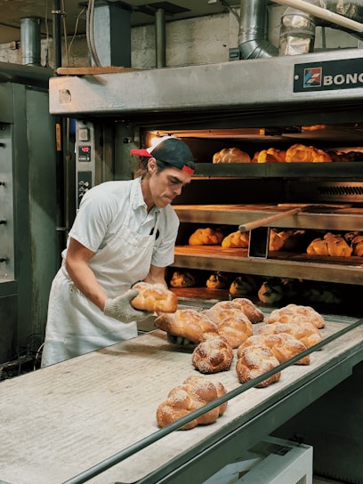 A baker wearing a white uniform and a red cap is carefully removing freshly baked bread from an industrial oven. The bread is displayed on a conveyor and appears golden brown with a dusting of flour on top. The setting is a commercial bakery with stainless steel equipment and machinery visible in the background.
