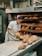A baker wearing a white uniform and a red cap is carefully removing freshly baked bread from an industrial oven. The bread is displayed on a conveyor and appears golden brown with a dusting of flour on top. The setting is a commercial bakery with stainless steel equipment and machinery visible in the background.