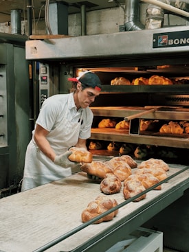 A baker wearing a white uniform and a red cap is carefully removing freshly baked bread from an industrial oven. The bread is displayed on a conveyor and appears golden brown with a dusting of flour on top. The setting is a commercial bakery with stainless steel equipment and machinery visible in the background.