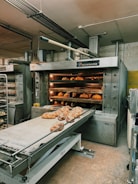 A commercial bakery setup featuring a large industrial oven with several racks of freshly baked bread. The oven has trays inside with bread visible through the open doors. There is also a conveyor belt in front with additional loaves placed on it.