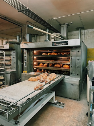A commercial bakery setup featuring a large industrial oven with several racks of freshly baked bread. The oven has trays inside with bread visible through the open doors. There is also a conveyor belt in front with additional loaves placed on it.