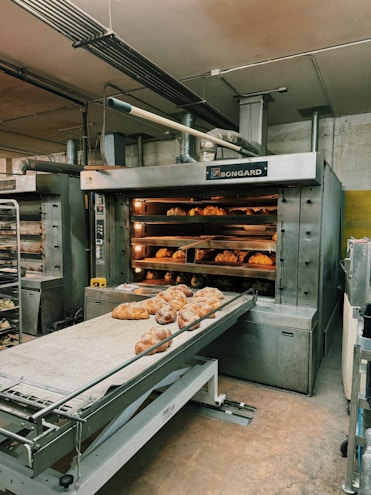 A commercial bakery setup featuring a large industrial oven with several racks of freshly baked bread. The oven has trays inside with bread visible through the open doors. There is also a conveyor belt in front with additional loaves placed on it.