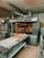 A commercial bakery setup featuring a large industrial oven with several racks of freshly baked bread. The oven has trays inside with bread visible through the open doors. There is also a conveyor belt in front with additional loaves placed on it.