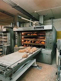 A commercial bakery setup featuring a large industrial oven with several racks of freshly baked bread. The oven has trays inside with bread visible through the open doors. There is also a conveyor belt in front with additional loaves placed on it.