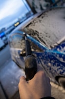 A close-up view of a person holding a pressure washer wand, spraying soapy water onto a car. The vehicle is covered in a layer of foam, and the scene takes place outdoors, possibly at an automatic car wash facility.