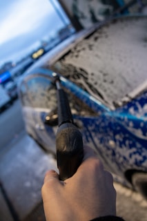 A close-up view of a person holding a pressure washer wand, spraying soapy water onto a car. The vehicle is covered in a layer of foam, and the scene takes place outdoors, possibly at an automatic car wash facility.
