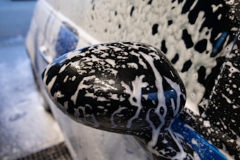 A close-up view of a car's side mirror covered entirely in soap suds, indicating it is being washed. The foamy bubbles create a pattern over the glossy black surface of the mirror, and the background shows part of the car also covered in soap. The scene suggests a car cleaning process in progress.
