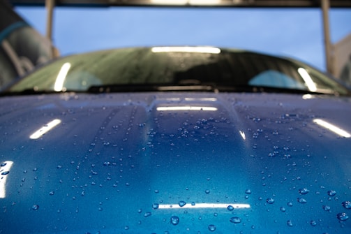 Close-up of a glossy black sports car hood with water droplets reflecting electric blue lighting.