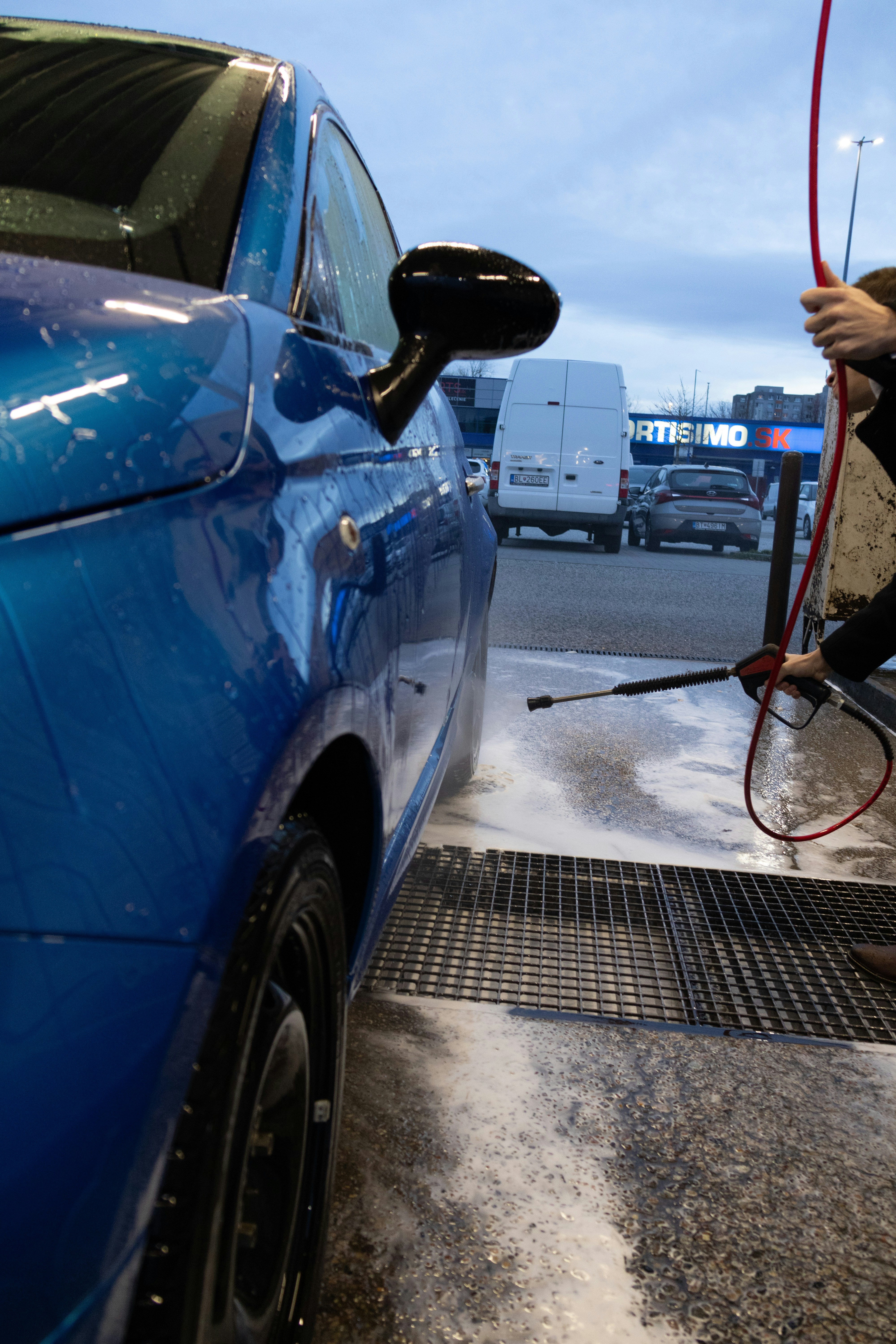 Car exterior being washed with water streams cascading over shiny blue vehicle surface in bright daylight