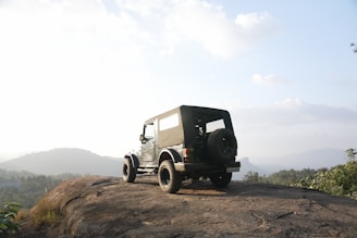 A rugged off-road vehicle is parked on a rocky ledge overlooking a scenic mountainous landscape. The background features rolling hills and a partly cloudy sky. The vehicle has a spare tire mounted on the back and appears to be positioned for an adventurous journey.