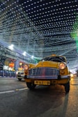 A yellow taxi is driving down a street at night, with a backdrop of a building decorated with bright, hanging lights forming a patterned canopy. There is another car partially visible in the background. The scene is illuminated by the decorative lights, creating a festive atmosphere.