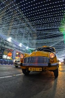 A yellow taxi is driving down a street at night, with a backdrop of a building decorated with bright, hanging lights forming a patterned canopy. There is another car partially visible in the background. The scene is illuminated by the decorative lights, creating a festive atmosphere.