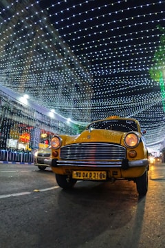 A yellow taxi is driving down a street at night, with a backdrop of a building decorated with bright, hanging lights forming a patterned canopy. There is another car partially visible in the background. The scene is illuminated by the decorative lights, creating a festive atmosphere.