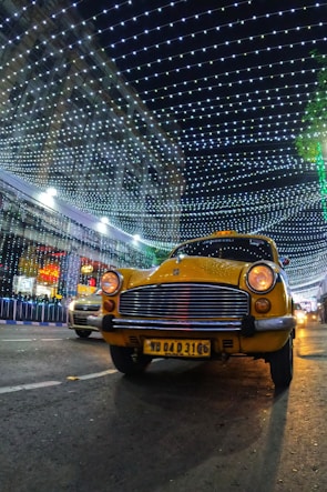A yellow taxi is driving down a street at night, with a backdrop of a building decorated with bright, hanging lights forming a patterned canopy. There is another car partially visible in the background. The scene is illuminated by the decorative lights, creating a festive atmosphere.